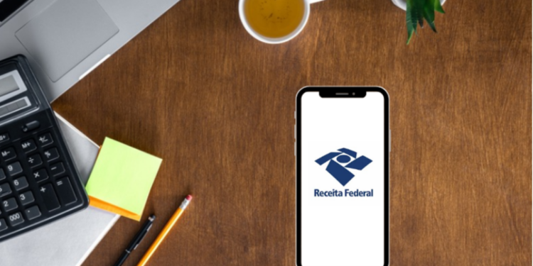 Mesa de escritório com celular mostrando a logo da Receita Federal.
