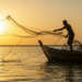 Pescador lança rede no mar ao pôr do sol em cenário sereno.