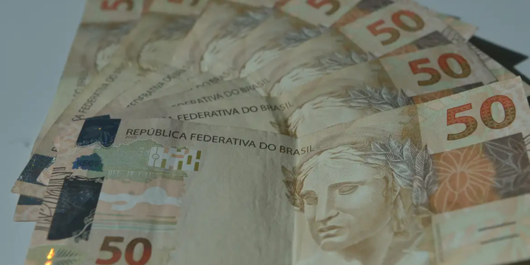 Imagem ilustrativa de cédulas de cinquenta reais espalhadas sobre superfície clara, em referência ao valor do Bolsa Família e aos adicionais que compõem a parcela de agosto