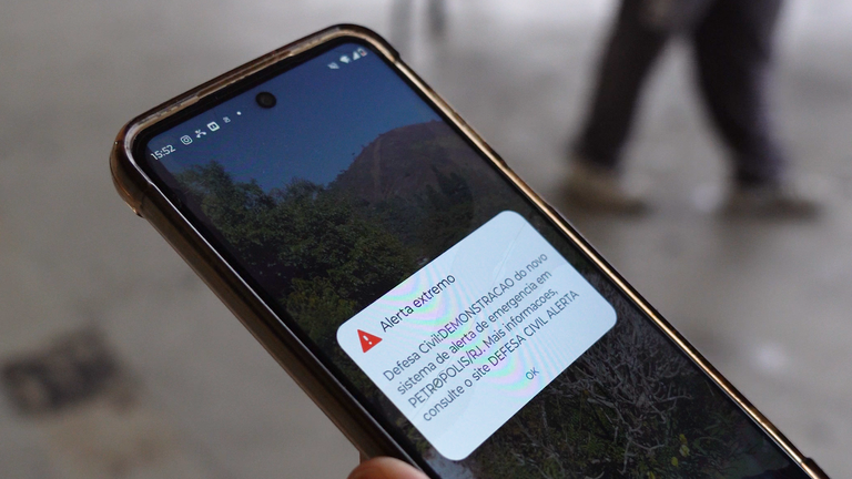 Celular na mão de uma pessoa exibindo notificação de alerta extremo da Defesa Civil sobre o novo sistema de alerta de emergência.