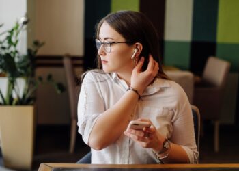 Imagem ilustrativa de mulher de óculos ajustando um fone sem fio enquanto consulta o smartphone em um café, remetendo à mulher que mantém o aparelho por perto para receber comunicados de segurança