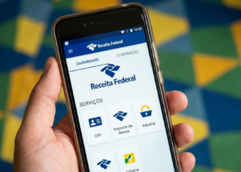 Imagem de mão segurando um celular com o aplicativo da Receita Federal aberto na tela de serviços, incluindo o Imposto de Renda