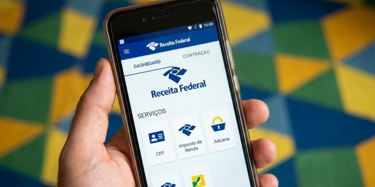 Imagem de mão segurando um celular com o aplicativo da Receita Federal aberto na tela de serviços, incluindo o Imposto de Renda