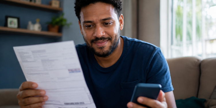 Imagem ilustrativa de homem sorrindo ao olhar um boleto em papel enquanto segura o celular na sala de casa, referência à renegociação de dívidas com juros menores