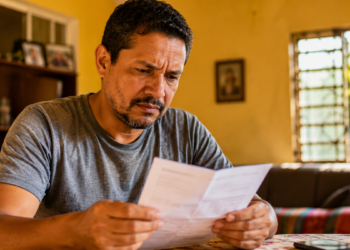 Imagem ilustrativa de homem lendo com atenção um papel dobrado sentado à mesa de casa, cena ligada à checagem de promessas de indenização que circulam pela internet.