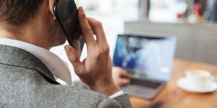 Imagem ilustrativa de homem visto de costas falando ao celular junto a um notebook em uma mesa, referência à identificação e ao bloqueio de números indesejados no próprio aparelho