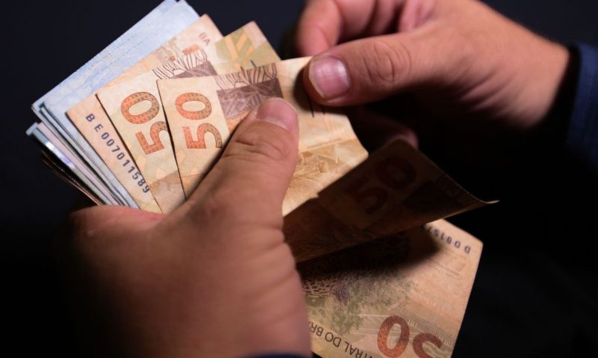 Até R$ 8.475,55 no INSS: depósitos de agosto serão feitos no início de setembro para beneficiários 2 Mãos contando um maço de cédulas de cinquenta reais sobre fundo escuro, referência aos valores pagos aos beneficiários conforme a data definida no calendário