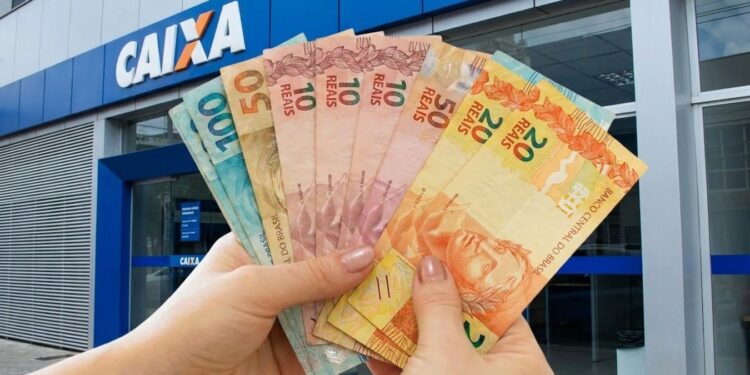 Imagem ilustrativa de mãos segurando cédulas de real em leque diante da fachada de uma agência da Caixa, em referência à liberação das parcelas do Pé-de-Meia
