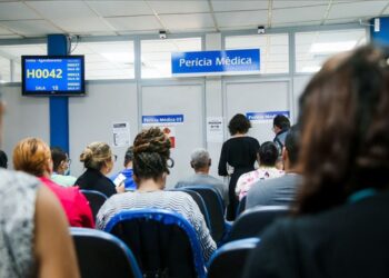 Sala de espera de uma agência do INSS com pessoas sentadas diante do painel de senhas e das salas identificadas como perícia médica.