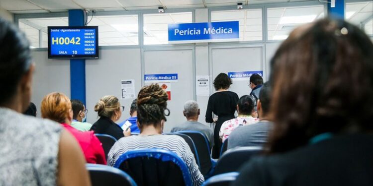 Sala de espera de uma agência do INSS com pessoas sentadas diante do painel de senhas e das salas identificadas como perícia médica.