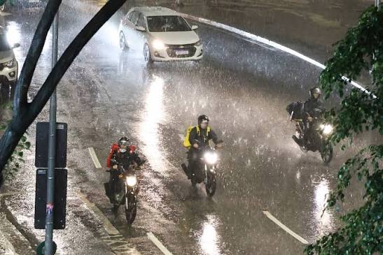 Imagem ilustrativa de motociclistas trafegando sob chuva intensa em uma avenida à noite, representando o impacto da chuva forte no trânsito