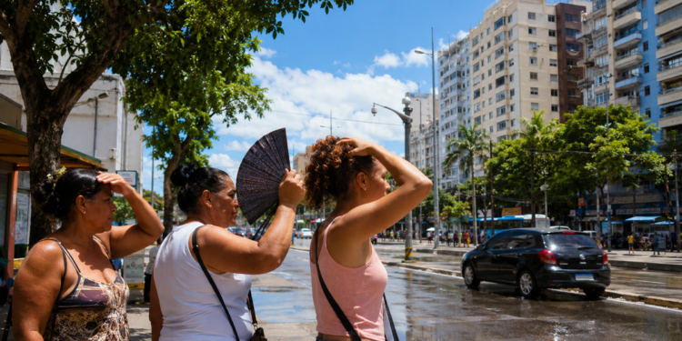 Imagem ilustrativa de pessoas se abanando e cobrindo a cabeça do sol forte em uma avenida arborizada de céu azul, referência ao contraste de clima entre as regiões durante a virada do tempo