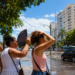 Imagem ilustrativa de pessoas se abanando e cobrindo a cabeça do sol forte em uma avenida arborizada de céu azul, referência ao contraste de clima entre as regiões durante a virada do tempo