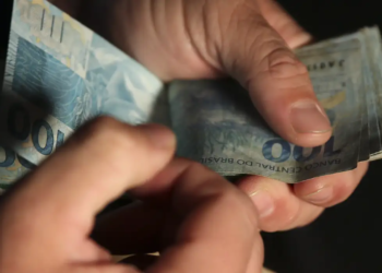 Imagem ilustrativa de mãos contando notas de cem reais sobre fundo escuro, tema da matéria sobre a estimativa do piso nacional para 2027.