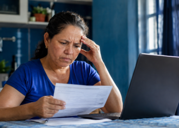 Imagem ilustrativa de mulher lendo um documento diante de um notebook, com a mão apoiada na cabeça, cena ligada à negativa de benefício do INSS tratada em projeto de lei.
