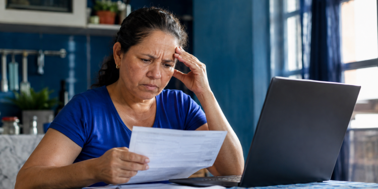 Imagem ilustrativa de mulher lendo um documento diante de um notebook, com a mão apoiada na cabeça, cena ligada à negativa de benefício do INSS tratada em projeto de lei.