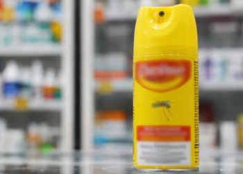 Imagem ilustrativa de frasco de spray repelente de insetos sobre um balcão, com prateleiras de farmácia ao fundo