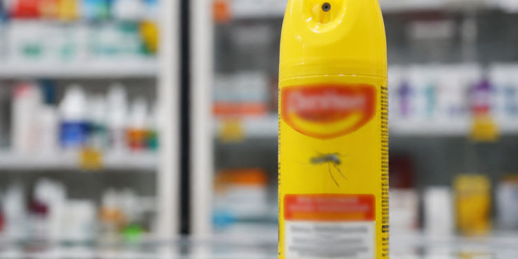 Imagem ilustrativa de frasco de spray repelente de insetos sobre um balcão, com prateleiras de farmácia ao fundo