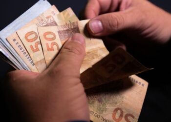 Imagem ilustrativa de mãos contando cédulas de cinquenta reais sobre fundo escuro, em referência à projeção do salário mínimo apresentada pelo governo para 2027