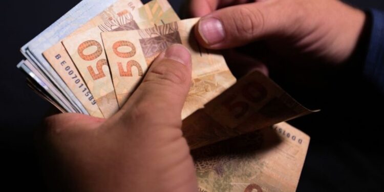 Imagem ilustrativa de mãos contando cédulas de cinquenta reais sobre fundo escuro, em referência à projeção do salário mínimo apresentada pelo governo para 2027