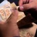Imagem ilustrativa de mãos contando cédulas de cinquenta reais sobre fundo escuro, em referência à projeção do salário mínimo apresentada pelo governo para 2027