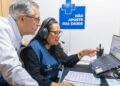 Atendente de headset e colete apontando para a tela de um notebook enquanto um homem acompanha ao lado, com o cartaz Não Aposte Sua Saúde ao fundo, referência ao apoio a pessoas afetadas por jogos e apostas