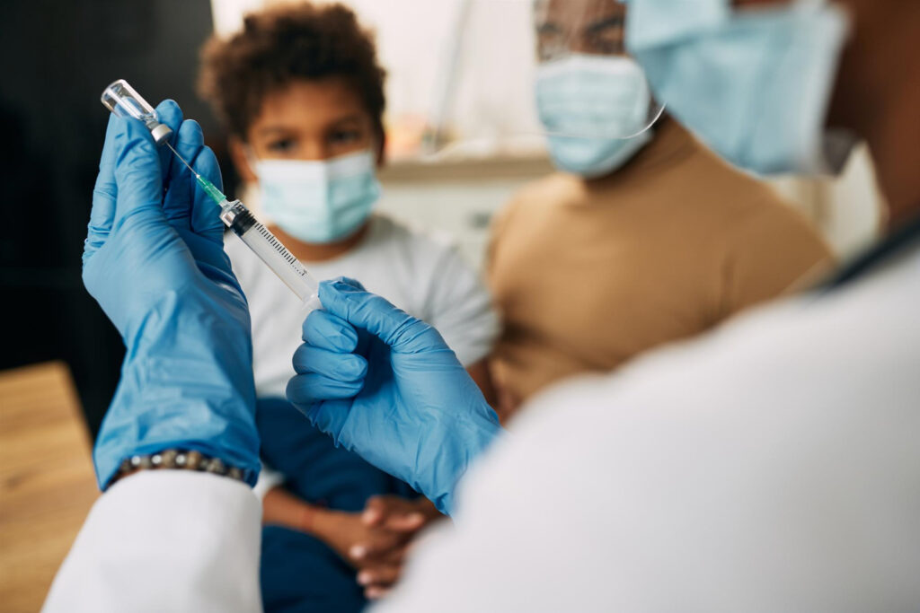Imagem ilustrativa de profissional de saúde preparando uma seringa com o frasco do imunizante diante de uma criança acompanhada por um adulto, em referência às vacinas disponíveis no SUS