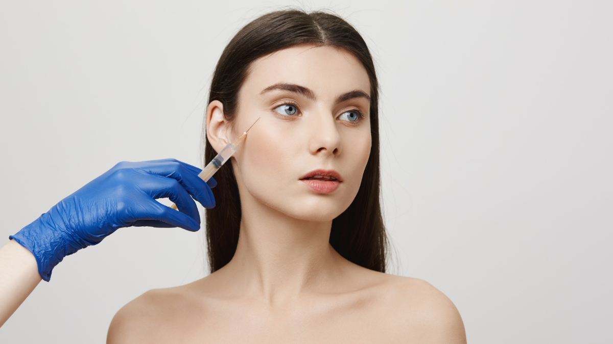 Mulher jovem recebe injeção de botox no rosto por profissional com luva azul em fundo claro.