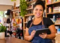 Empreendedora sorri atrás do balcão de sua loja de bairro enquanto registra vendas em um tablet.