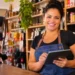 Empreendedora sorri atrás do balcão de sua loja de bairro enquanto registra vendas em um tablet.