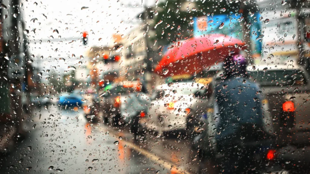 Chuva intensa cobre uma rua movimentada, com gotas de água no vidro e veículos circulando sob o tempo chuvoso.