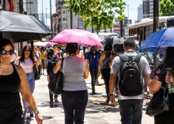 Pessoas caminham por uma rua movimentada sob sol intenso, usando guarda-chuvas para se proteger do forte calor.