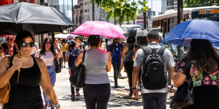 Pessoas caminham por uma rua movimentada sob sol intenso, usando guarda-chuvas para se proteger do forte calor.