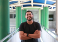 Estudante sorridente no corredor da faculdade com mochila e camiseta preta.