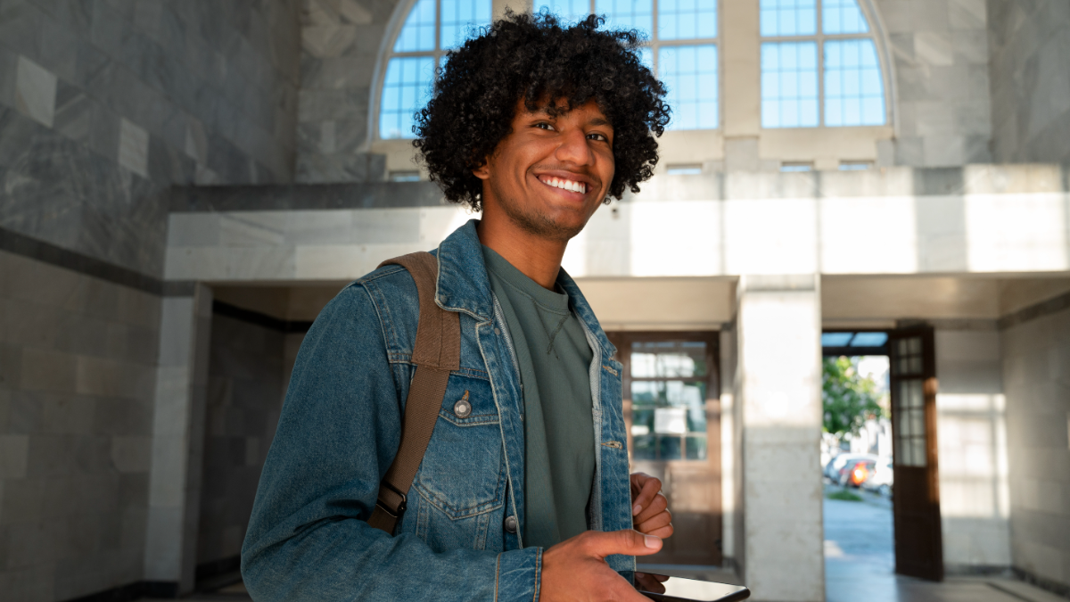 Jovem sorridente com mochila na entrada de prédio iluminado.