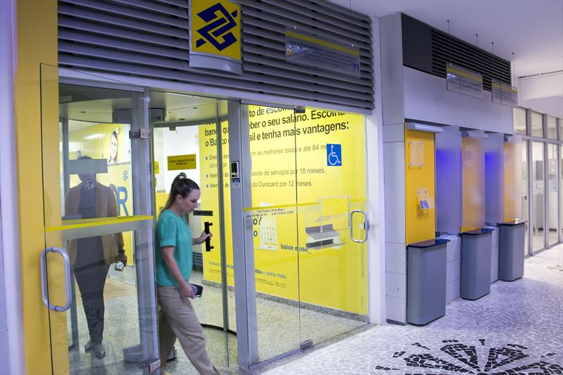 Mulher saindo de agência Banco do Brasil, fachada amarela, caixas eletrônicos ao lado.