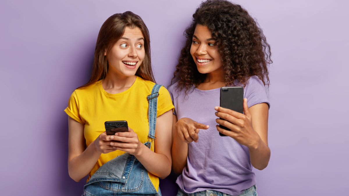 Amigas se divertem compartilhando algo no celular com expressões alegres.
