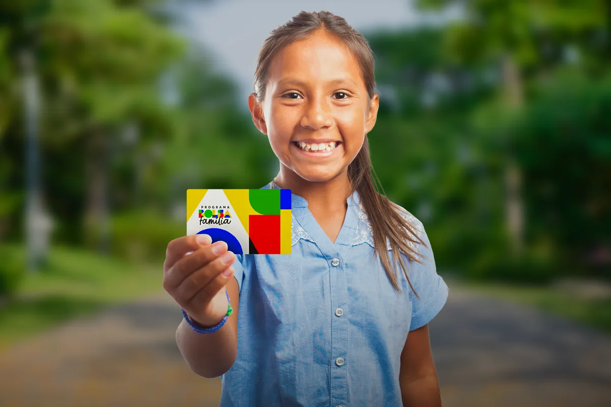 Menina sorridente exibe cartão do Bolsa Família ao ar livre.