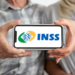 Duas pessoas seguram celular exibindo logo do INSS, foco nas mãos e aparelho.