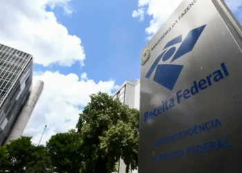 Placa da Receita Federal em frente a prédio moderno cercado de árvores sob céu azul com nuvens.