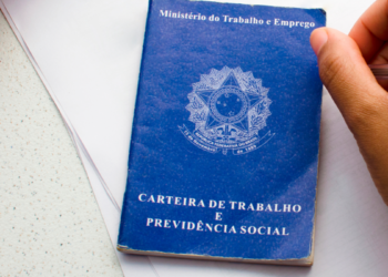 Mão segurando caneta próxima à Carteira de Trabalho e Previdência Social azul.