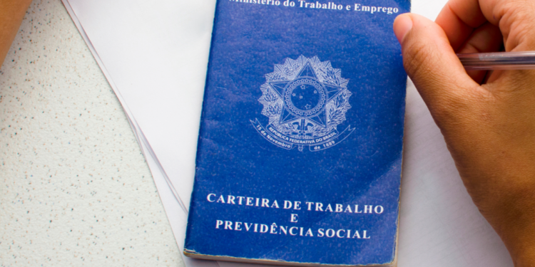 Mão segurando caneta próxima à Carteira de Trabalho e Previdência Social azul.
