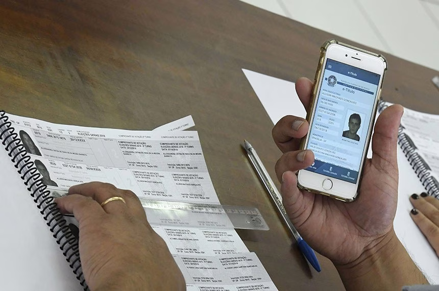 Eleitor segurando celular com o aplicativo e-Título aberto, com foto e dados, ao lado do caderno de votação com comprovantes sobre a mesa da seção eleitoral