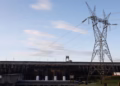 Barragem de usina hidrelétrica com torre de transmissão de energia em primeiro plano, sob céu azul, em referência à conta de luz de outubro