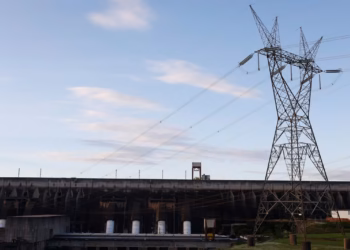Barragem de usina hidrelétrica com torre de transmissão de energia em primeiro plano, sob céu azul, em referência à conta de luz de outubro