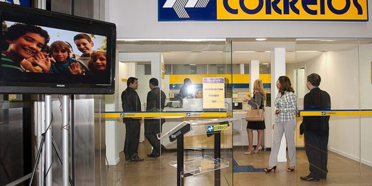 Clientes e funcionários dentro de uma agência dos Correios, com o letreiro da estatal acima dos guichês de atendimento.