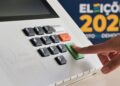 Imagem ilustrativa: dedo aperta a tecla confirma da urna eletrônica em arte das Eleições 2026, sobre o voto facultativo