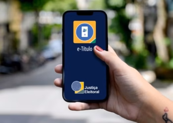 Imagem ilustrativa de mão segurando celular com a tela de abertura do aplicativo e-Título, da Justiça Eleitoral, em uma rua arborizada