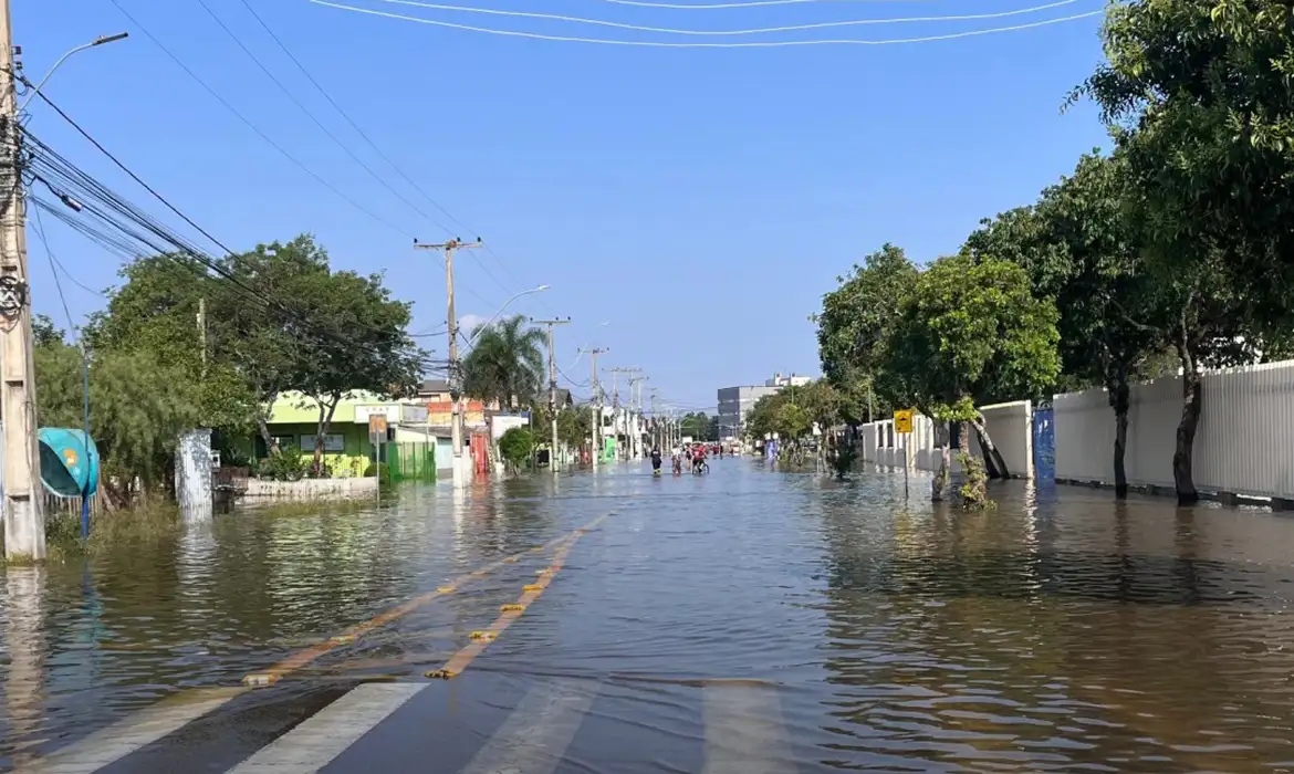 Rua residencial completamente alagada após temporal, com a água cobrindo o asfalto e moradores ao fundo em meio à enchente.