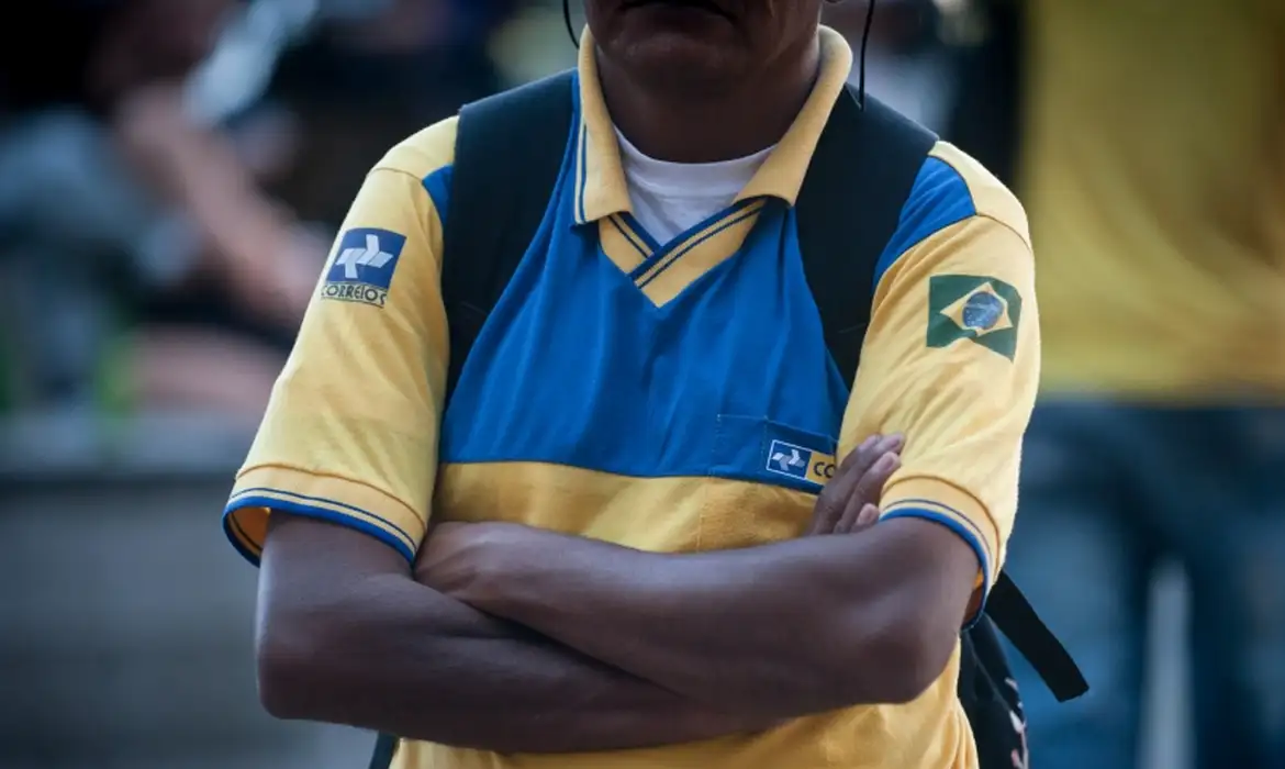 Imagem ilustrativa: carteiro de uniforme amarelo e azul dos Correios com os braços cruzados, em referência à greve dos Correios